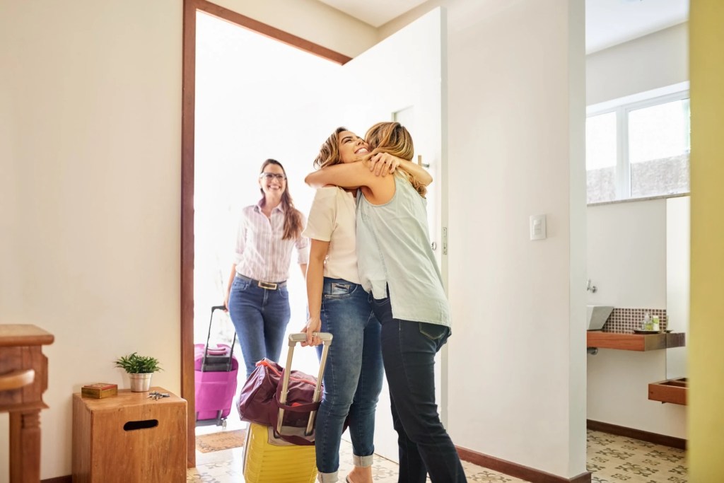 Two women hugging in a home entryway as a third arrives with suitcases