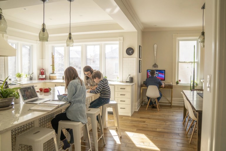 Woman helping two children with schoolwork at a kitchen island while a man works nearby