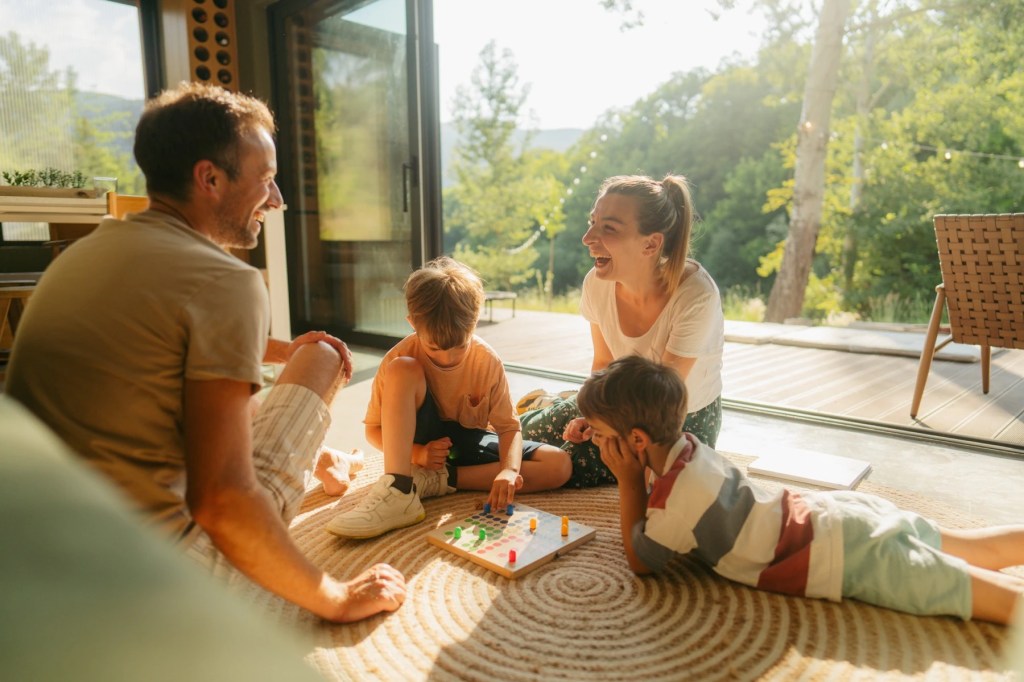 Family of four laughing over a board game on the floor beside open garden doors
