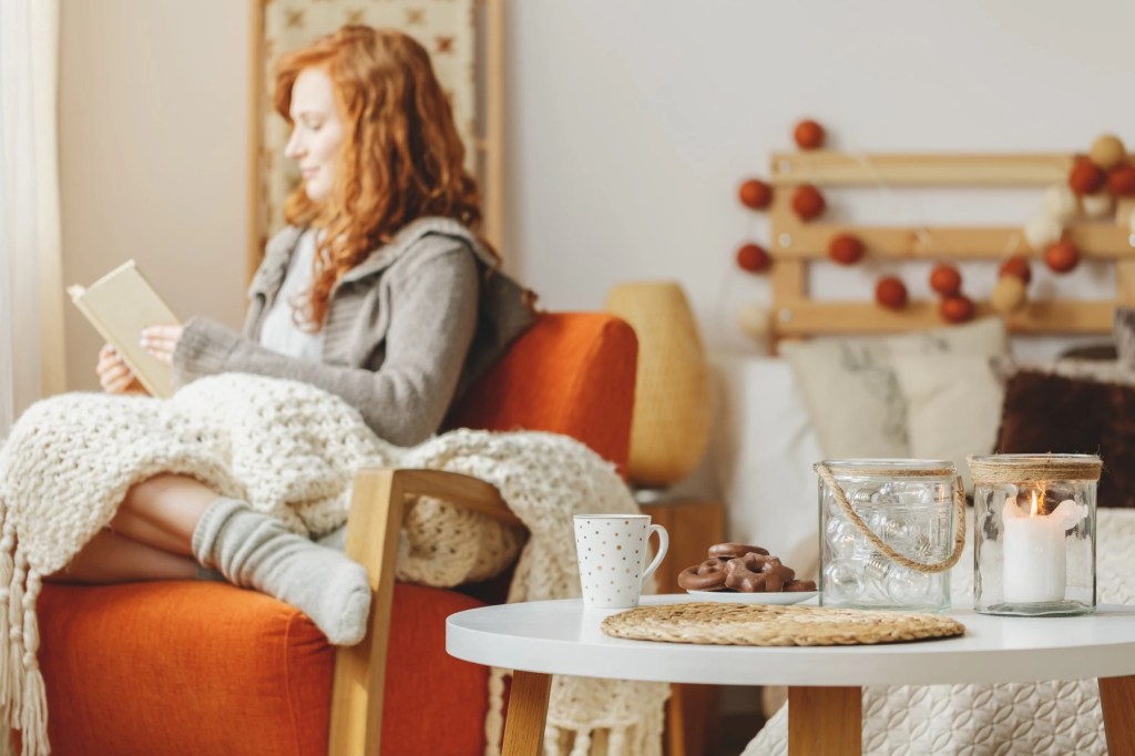 Woman reading in an armchair under a knitted blanket beside a lit candle