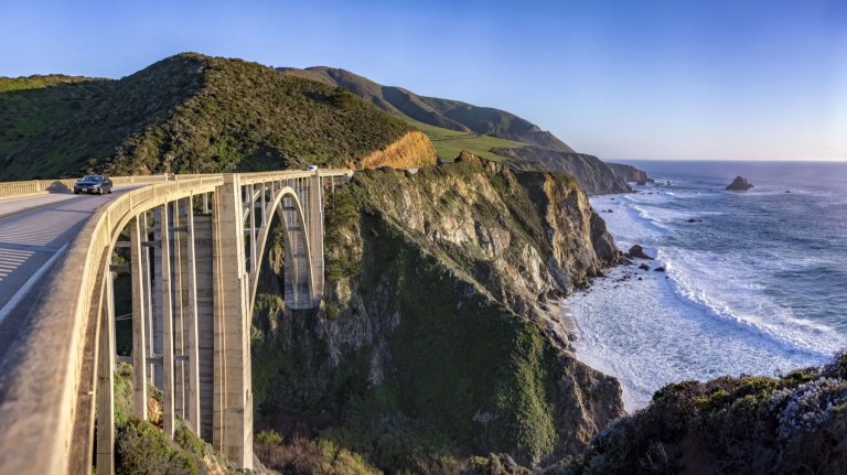 A car crossing a tall concrete arch bridge on a coastal cliff road