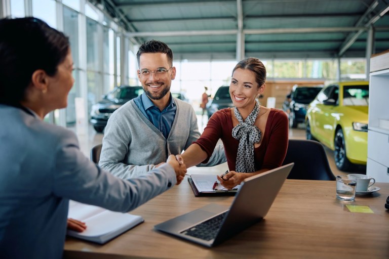 Couple shaking hands with a saleswoman at a car dealership desk