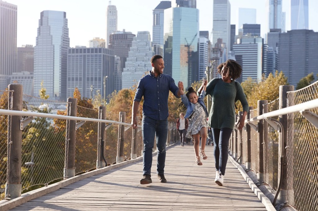 Family walking hand in hand across a footbridge with a city skyline behind