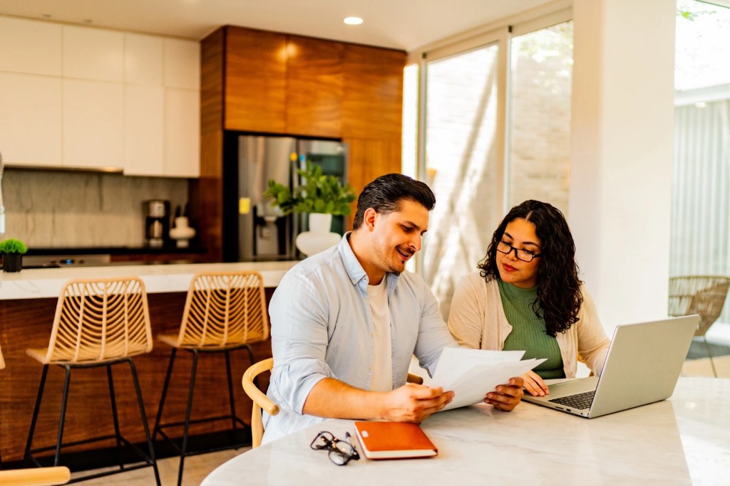 Man and woman reviewing paperwork at a kitchen table with a laptop
