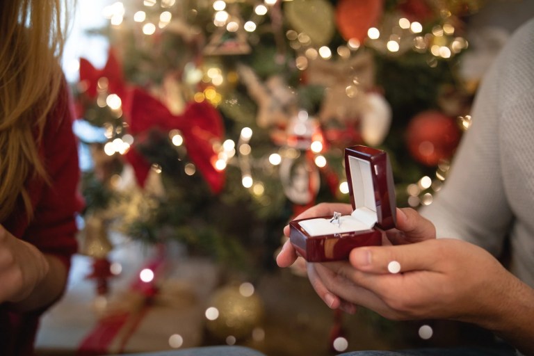 Person holding an open ring box in front of a lit Christmas tree